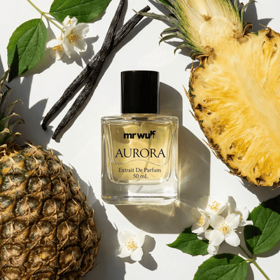 Aurora Cologne bottle surrounded by pineapple, vanilla sticks, and flowers, showcasing fresh citrus–aromatic fragrance.