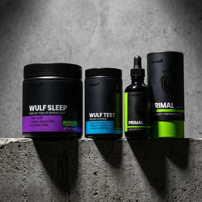 Three supplement containers from the brand Mr Wulf are lined up on a textured gray concrete ledge under dramatic spotlighting. The products include Wulf Sleep, Wulf Test and Primal, each in black packaging with distinct accent colors (purple, blue, green, and black/green).